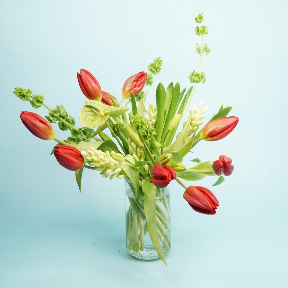 A bouquet of red tulips and green foliage in a clear glass vase against a light blue background, conveying a fresh and vibrant atmosphere.