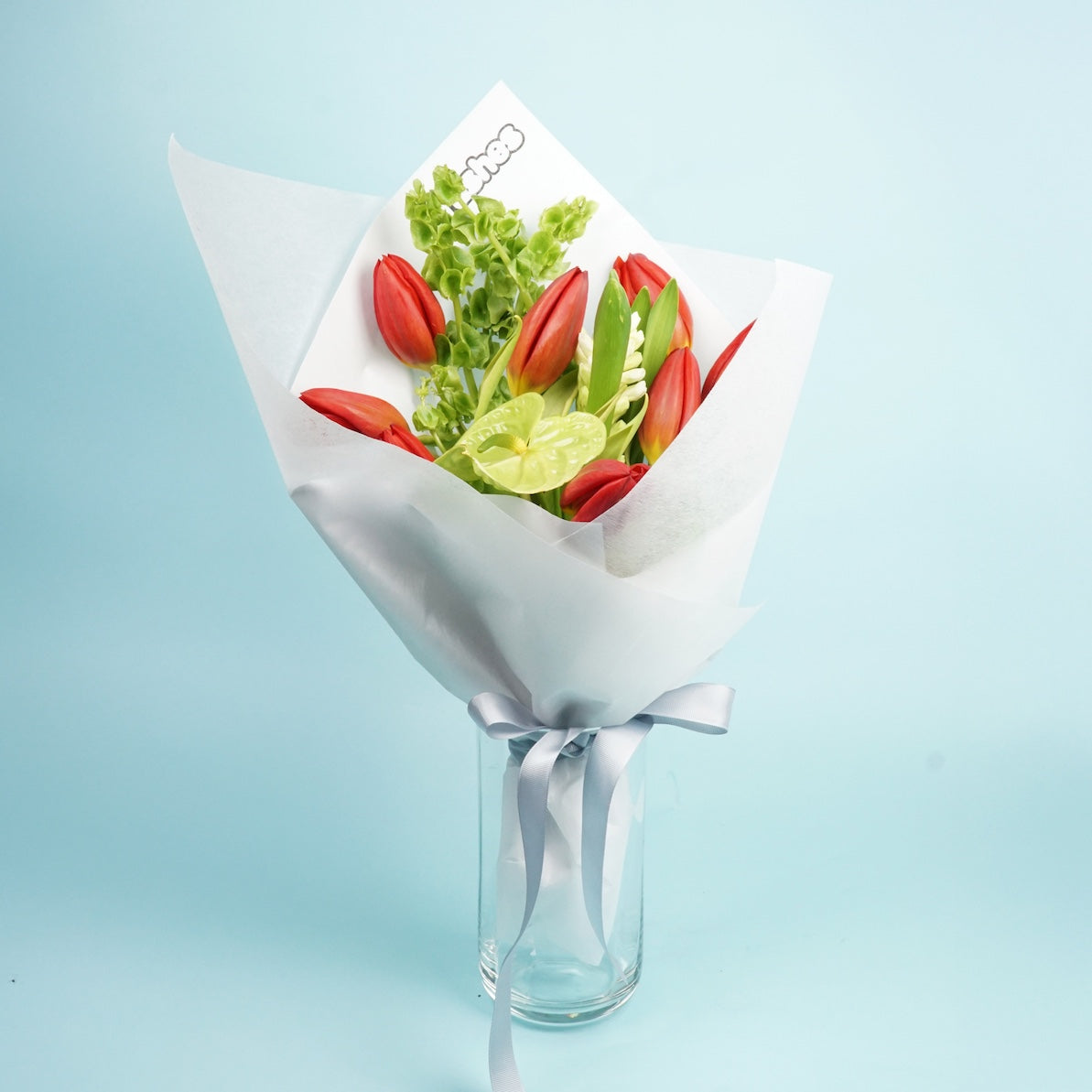 A bouquet of red tulips and green flora wrapped in white paper with a light blue ribbon, set in a glass vase against a soft blue background.