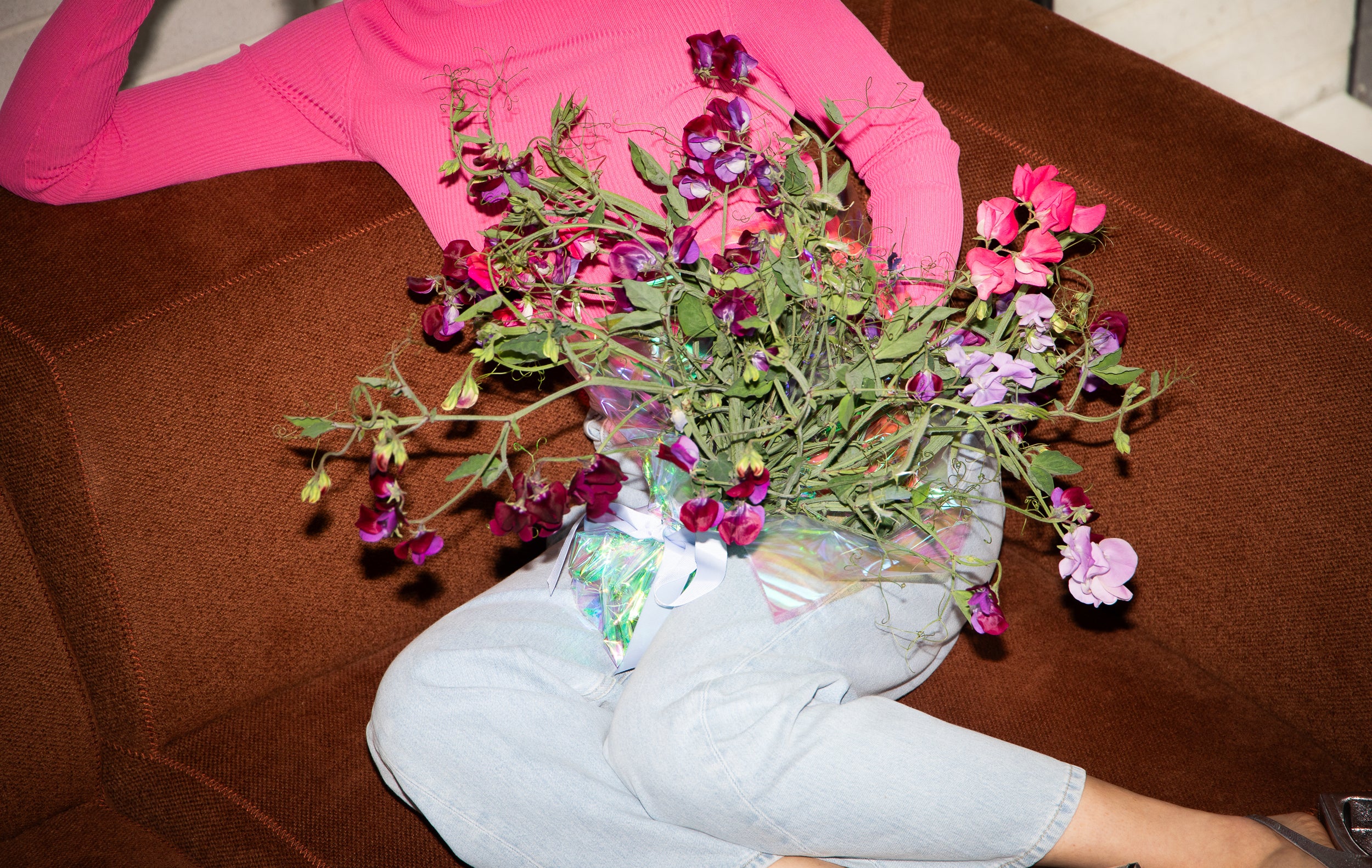 Person in a bright pink sweater and light jeans lounges on a brown sofa, holding a vibrant bouquet of purple and pink flowers, creating a playful, relaxed vibe.