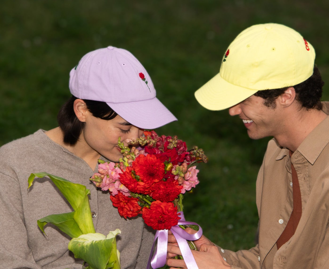 A person in a purple cap smells a vibrant bouquet of red and pink flowers, while another in a yellow cap smiles warmly. The scene suggests a joyful, intimate moment outdoors.