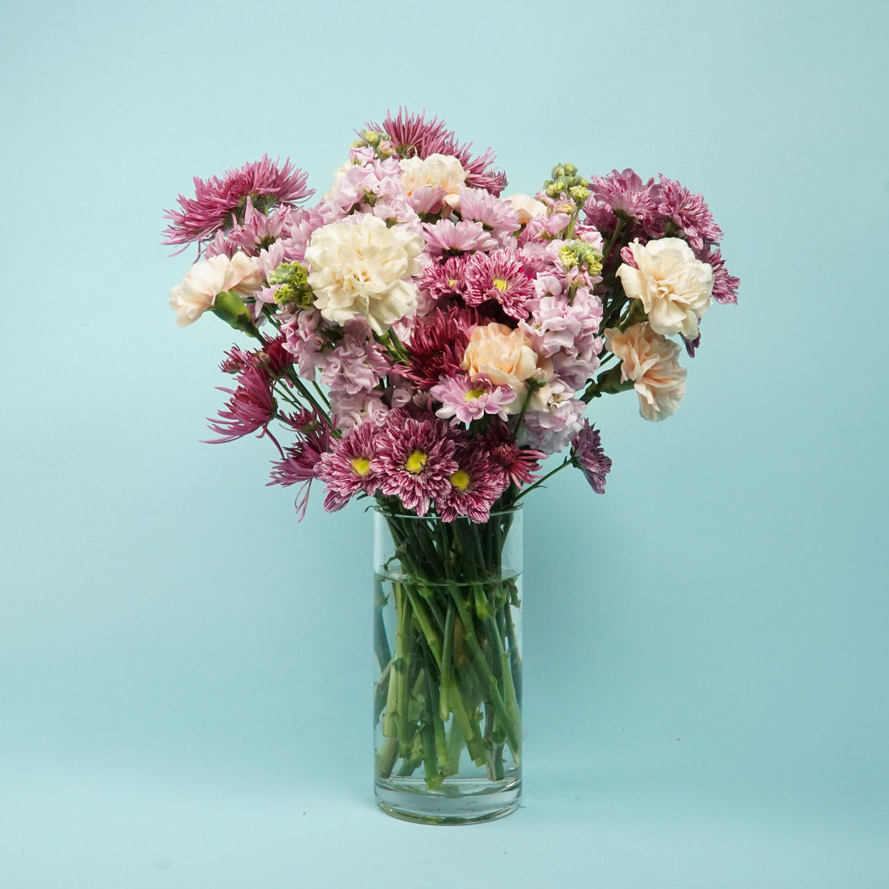 A vibrant bouquet of pink, purple, and cream flowers in a clear glass vase against a light blue background. The arrangement is lush and cheerful.