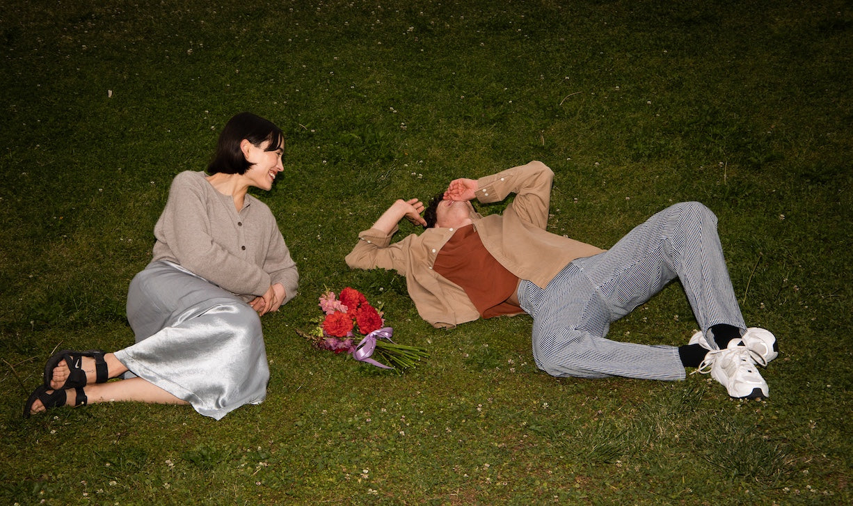 A couple relaxes on lush green grass, smiling warmly at each other. A bouquet of red and pink flowers lies between them, conveying a sense of joy and intimacy.